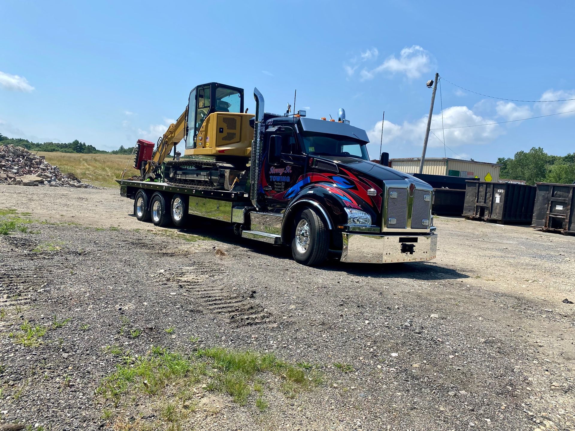 Red and blue heavy-duty tow truck carrying a yellow excavator in a gravel lot under a clear sky.