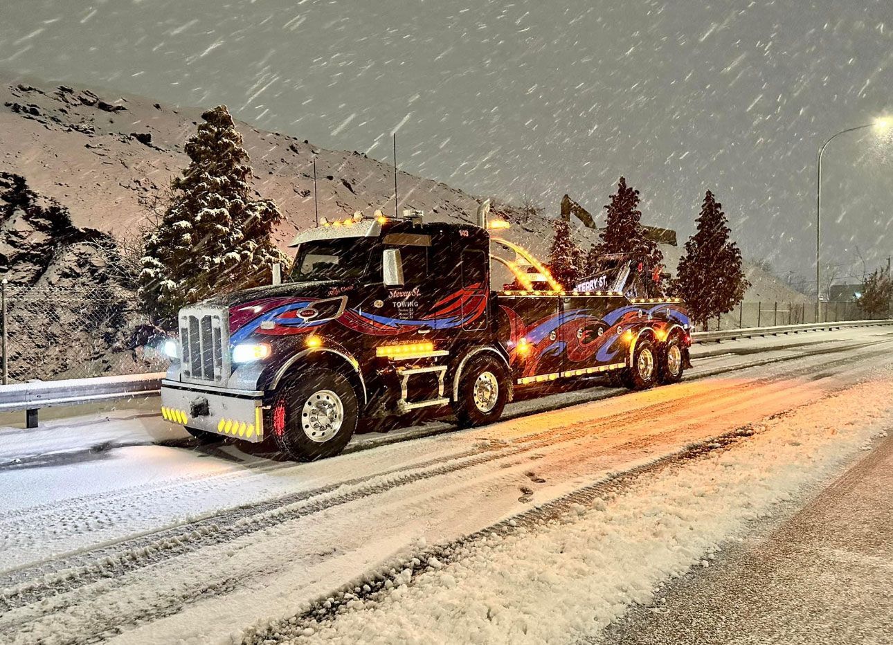 Tow truck hauling a car on a snowy highway at night, with headlights and flashing lights in snowfall.