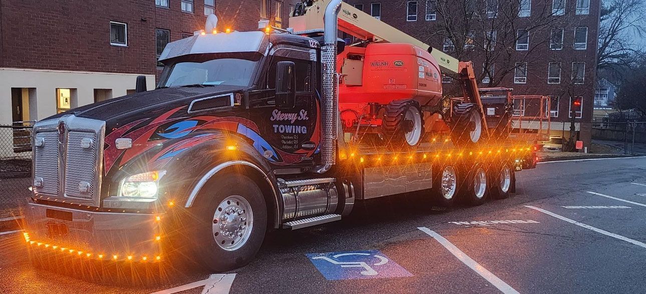 Black tow truck hauling a car on a rainy city street at dusk, lights glowing orange.