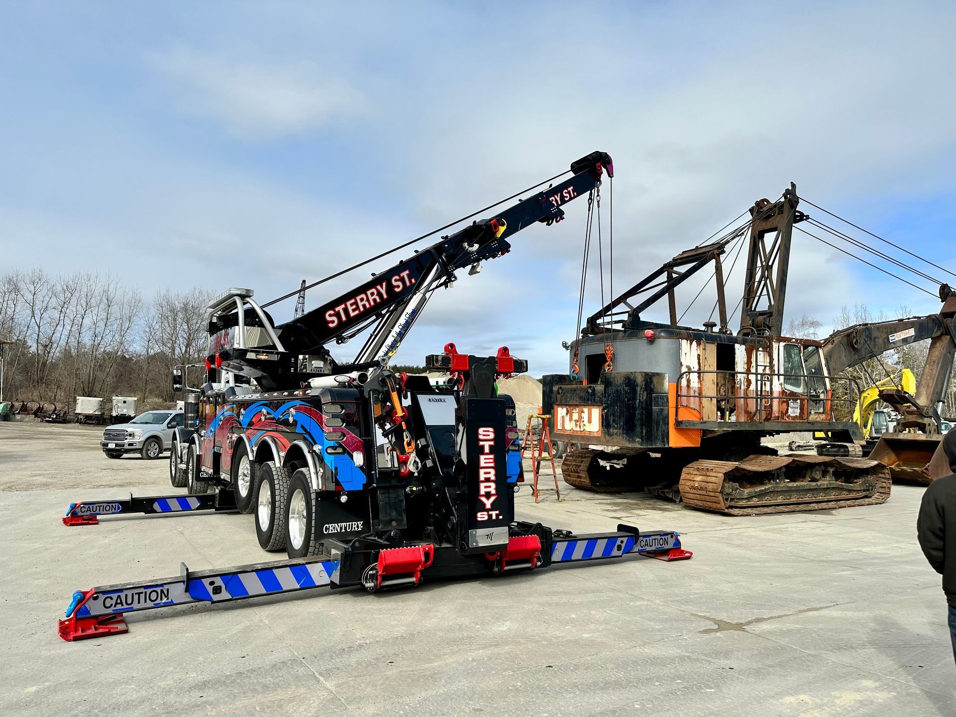 Black-and-red tow truck with extended boom parked beside a damaged excavator on a gravel lot.