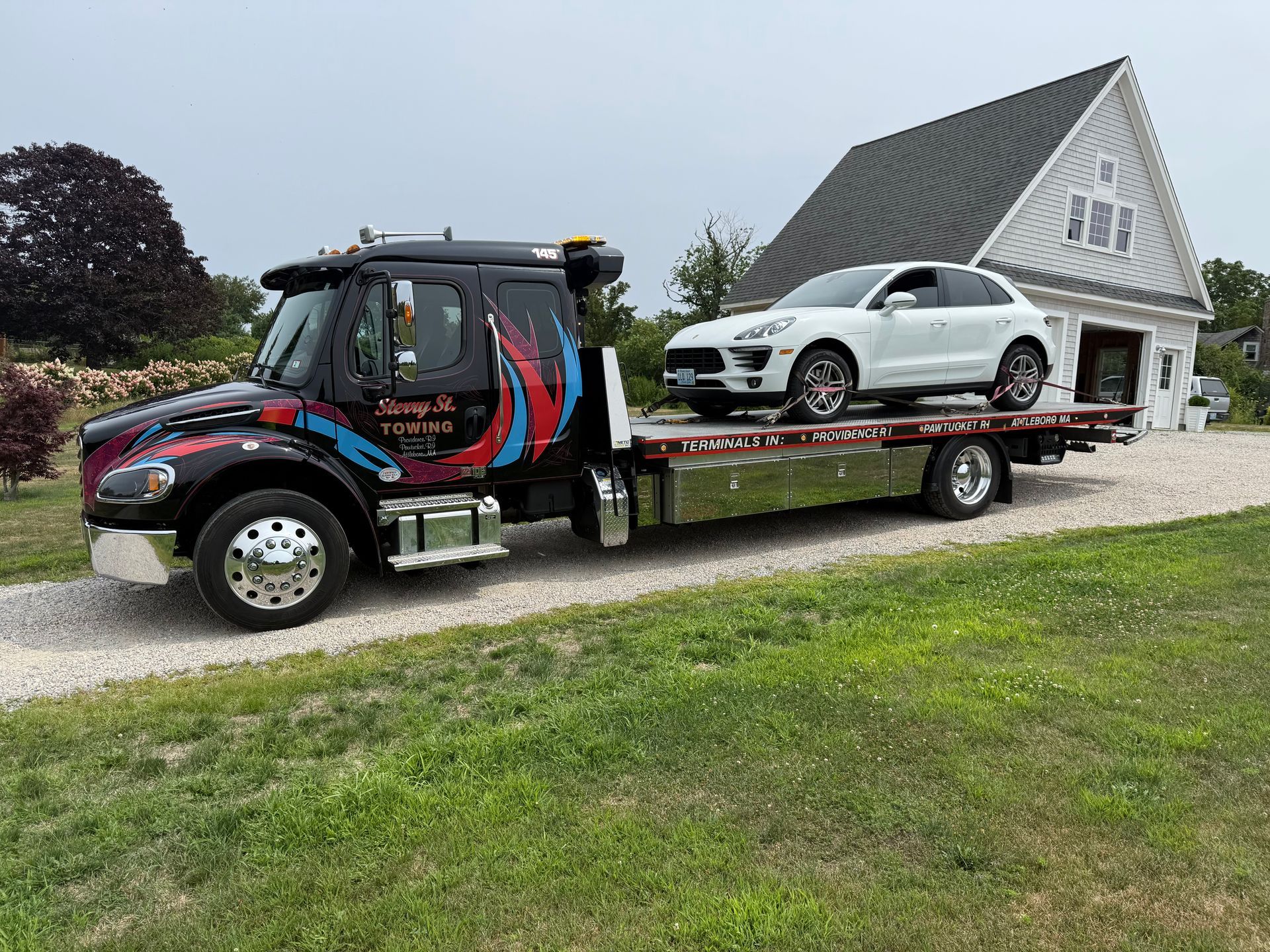 Tow truck carrying a white sports car on a flatbed beside a white house on a gravel driveway