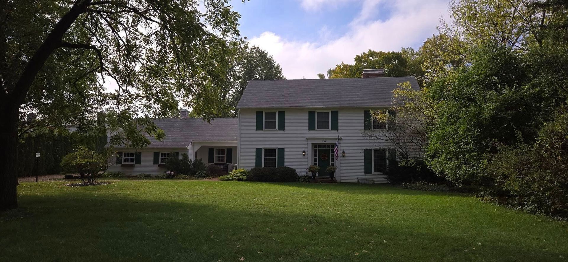 White two-story house with green shutters, in a grassy yard, trees surround the building.