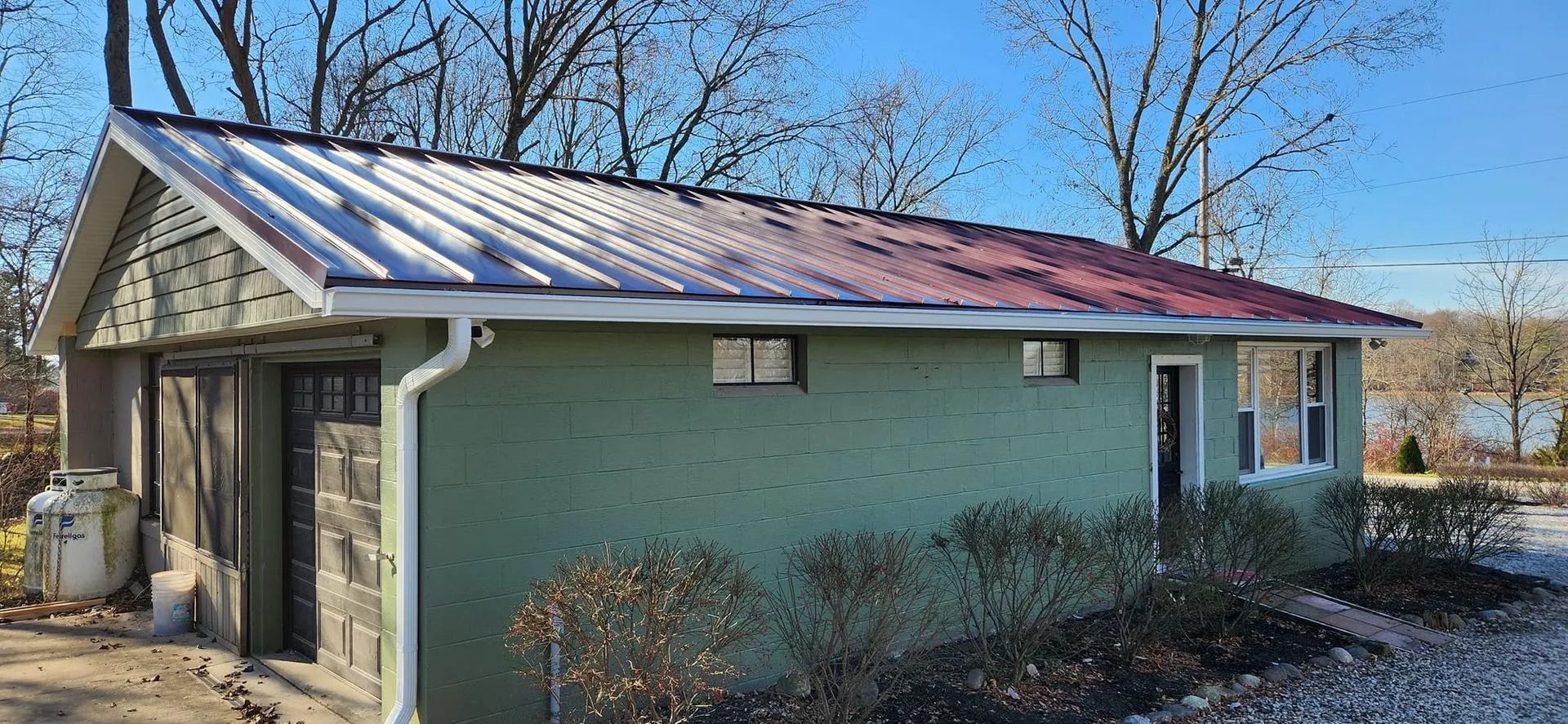 A green building with a metal roof, a garage, and a small garden in front. Trees and water are visible.