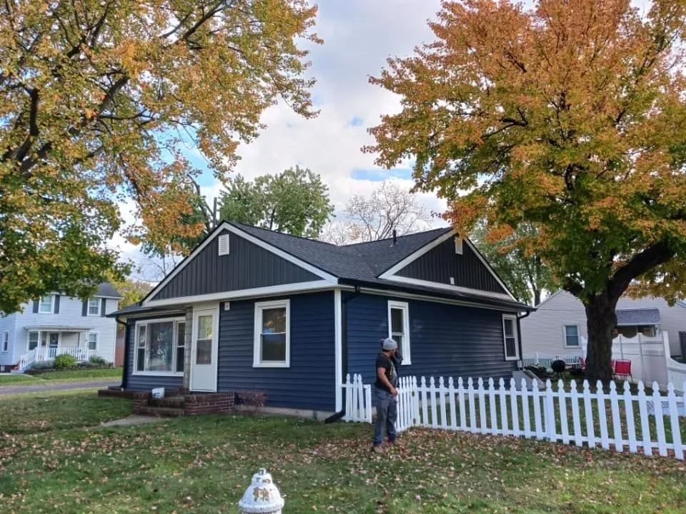 Blue house with white picket fence and trees with fall foliage. Person standing.