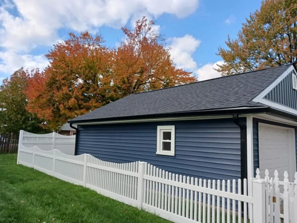 Blue-sided garage with white trim and roof, white picket fence, and colorful fall trees under a blue sky.