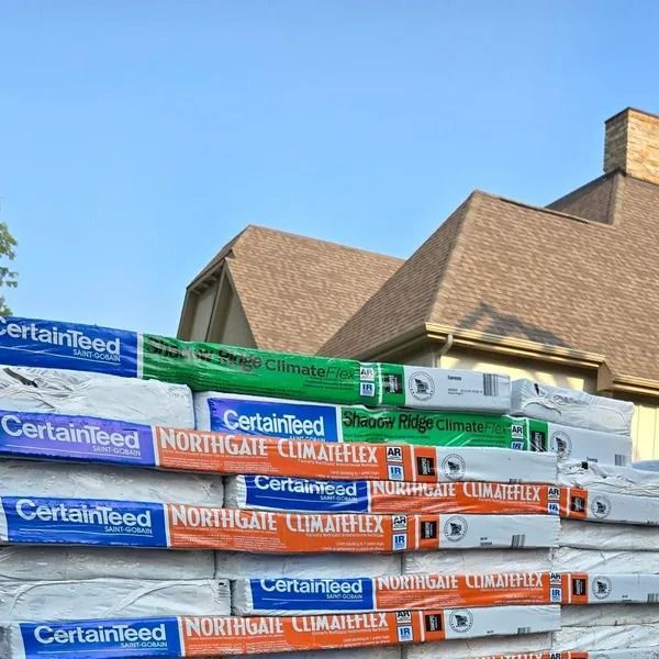 Piles of CertainTeed roofing materials in front of a house with a brown roof and blue sky.