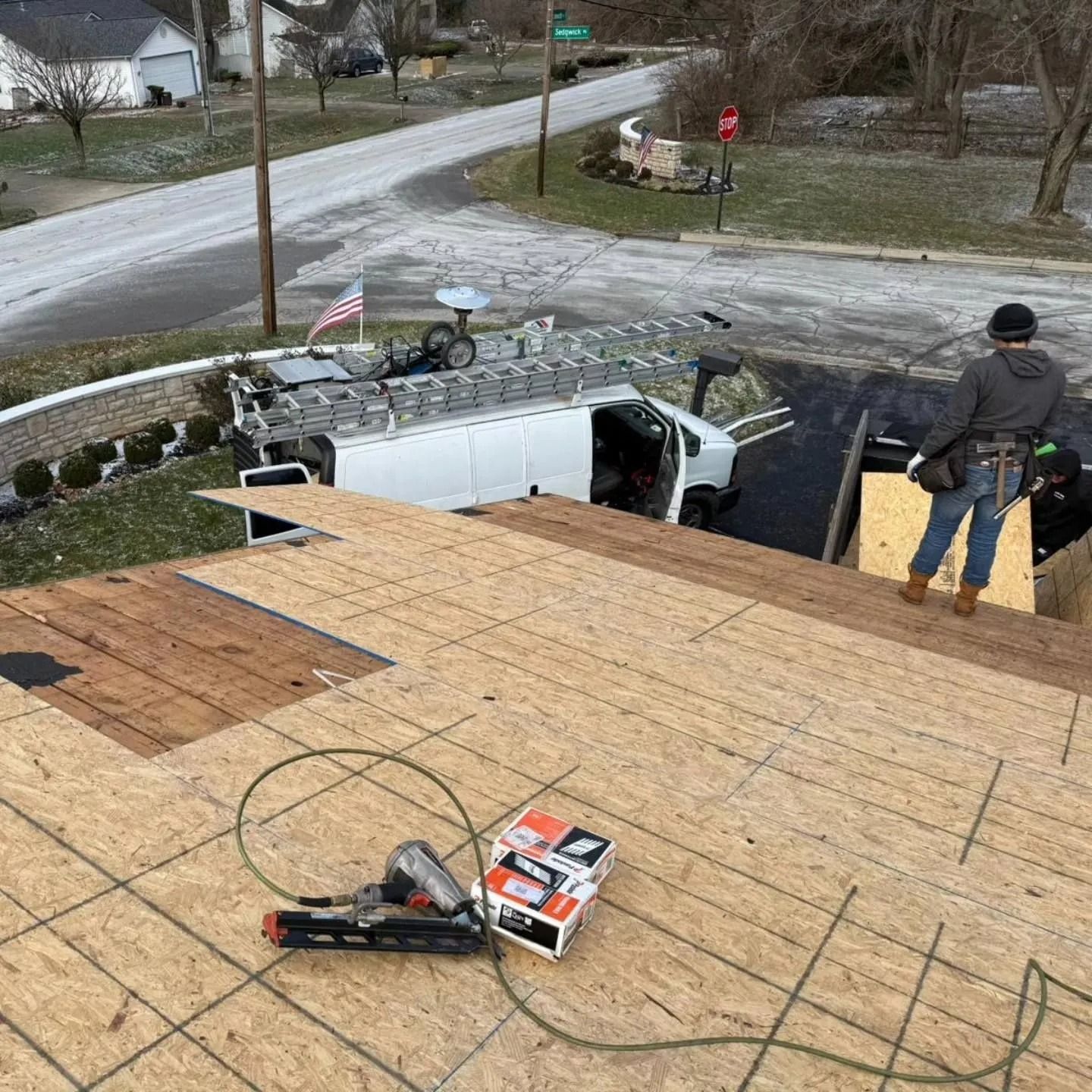 Roofer on roof with supplies, white van with ladder parked near road. American flag visible.
