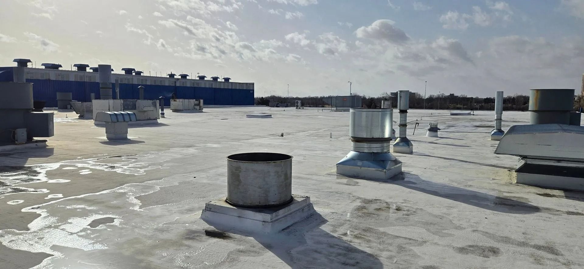 View of a white flat roof with metal vents and a building in the background under a cloudy sky.