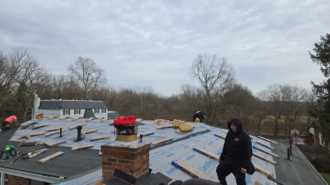Workers on a roof install roofing materials, cloudy day. Brick chimney, bare trees in the background.