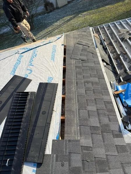 Roofing work: installing shingles on a home. Black shingles, blue underlayment, person standing nearby.