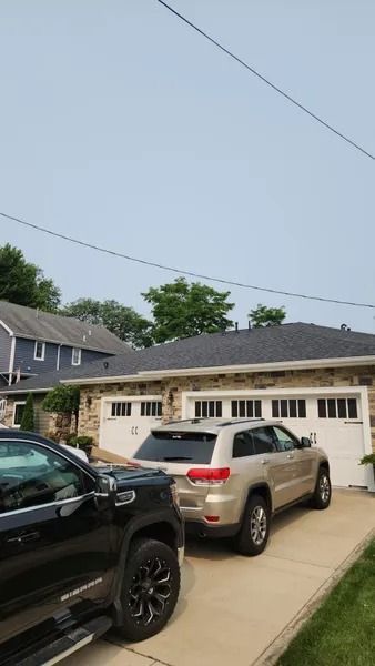 Two cars parked in a driveway in front of a garage. Houses and a blue sky are in the background.