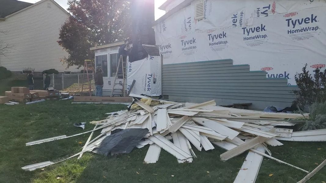 Construction site: debris pile in front of house with Tyvek wrap, lumber, and worker visible.