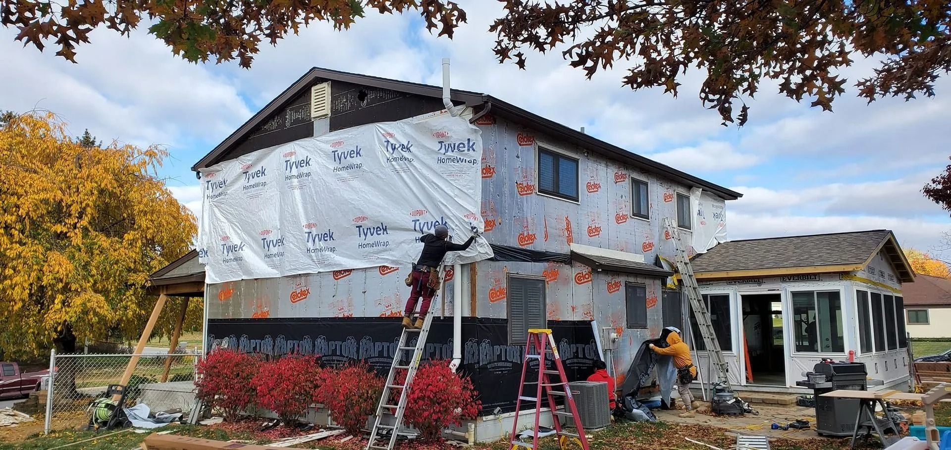 House under construction, workers installing siding, surrounded by foliage.