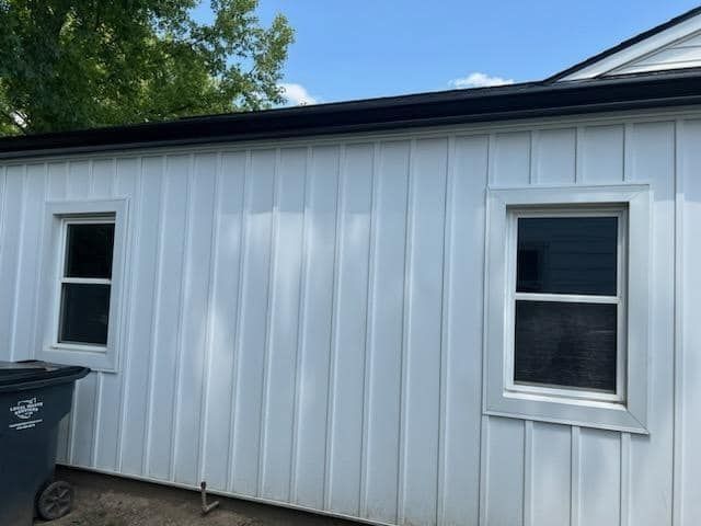 White vertical siding on a building with two windows and a black gutter under a blue sky.