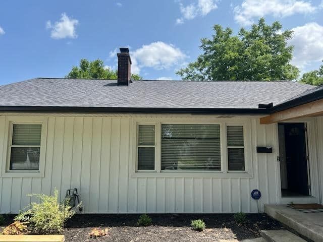 White house with black trim and roof. Chimney on the roof, trees in the background.
