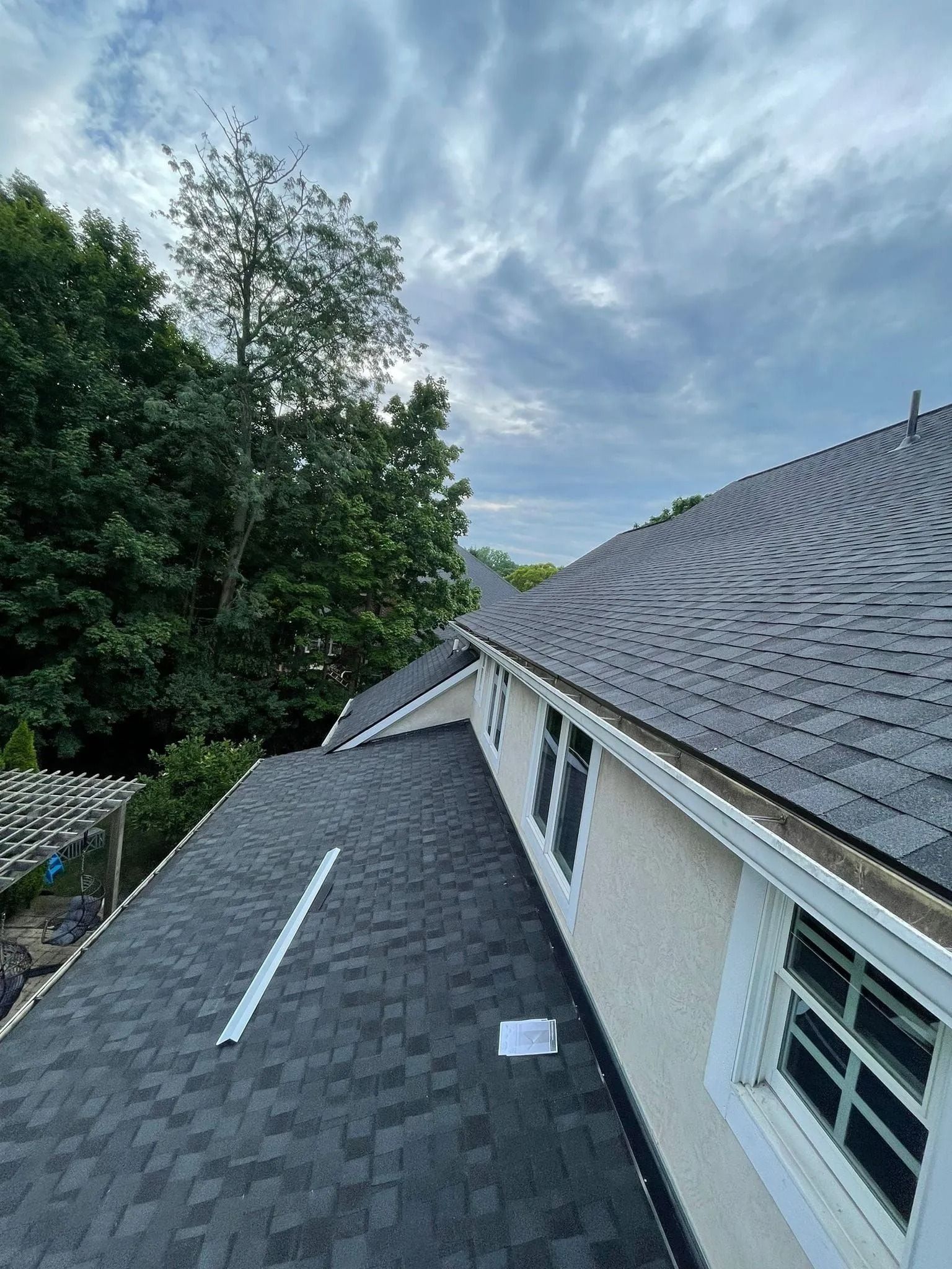 Rooftop view of a house with dark shingles, white trim, and windows, trees, and a cloudy sky.