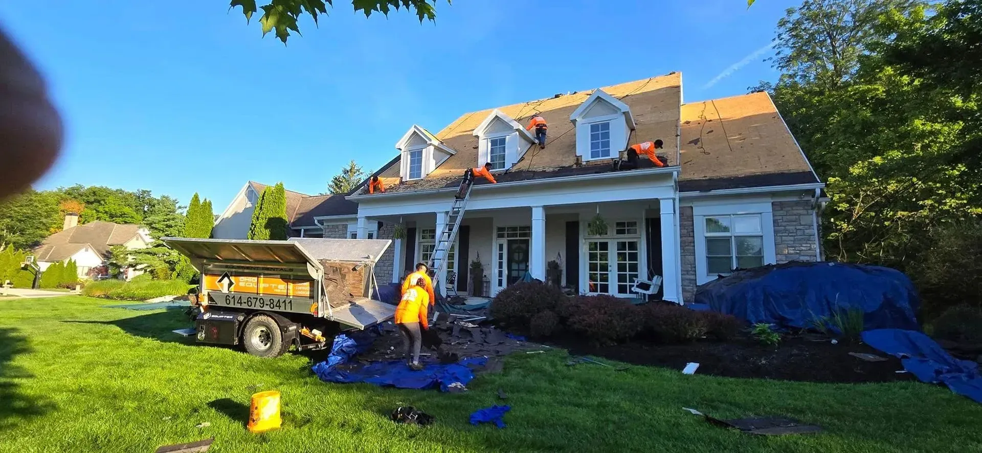 Roofers working on a house with blue tarp, a truck, and green lawn.
