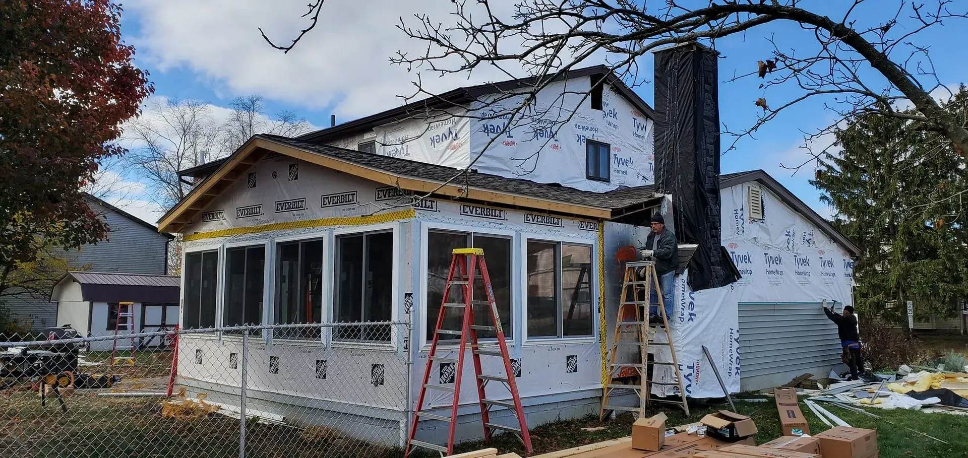 House under construction with a sunroom addition. Workers on ladders near a chimney and garage.