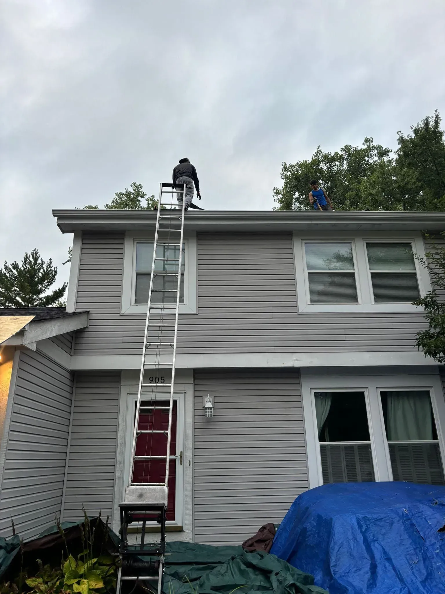 Two people on a roof cleaning gutters; one is high on a ladder. Grey siding, cloudy sky.