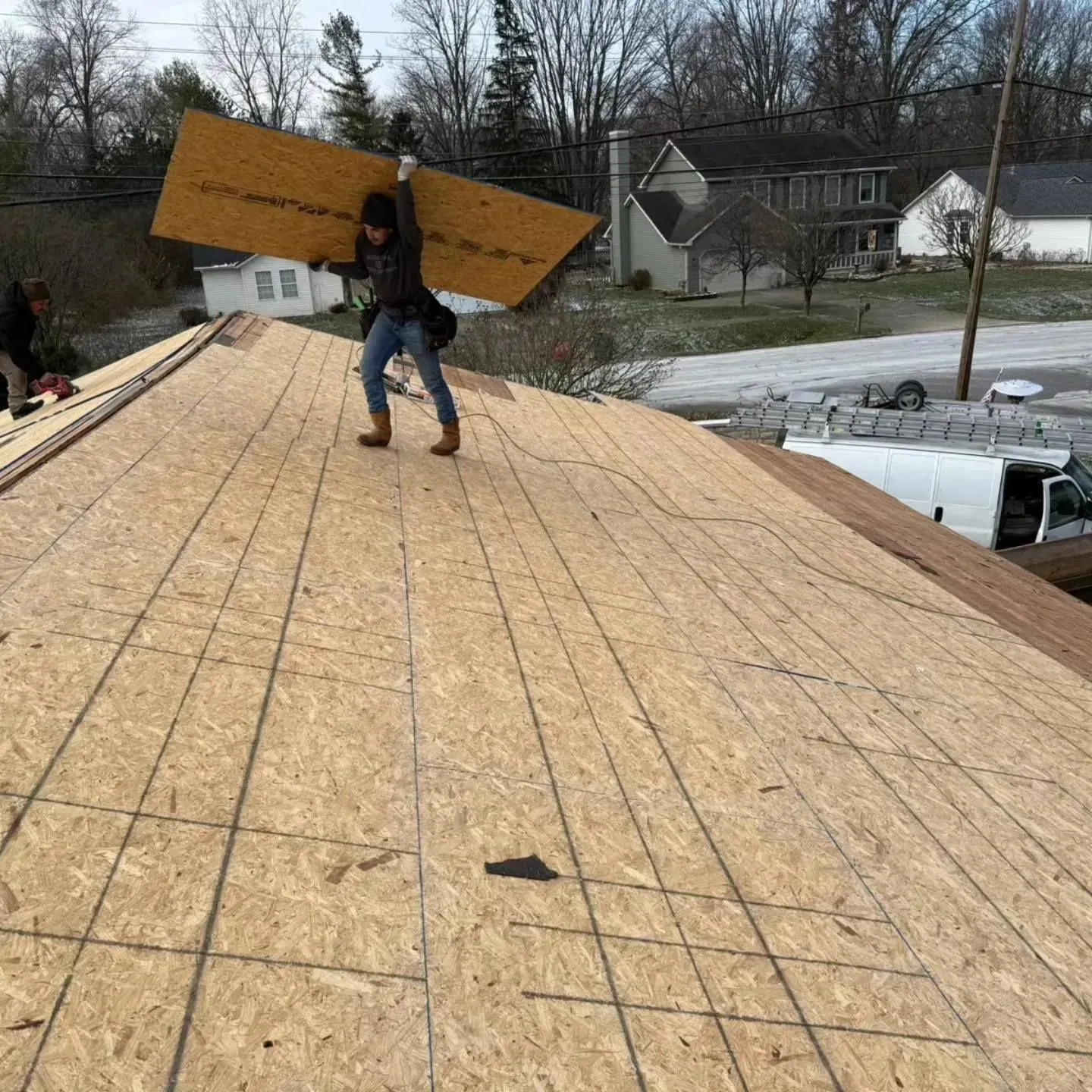 Roofer carrying large wood panel on a roof with completed underlayment. Houses and a van are in the background.