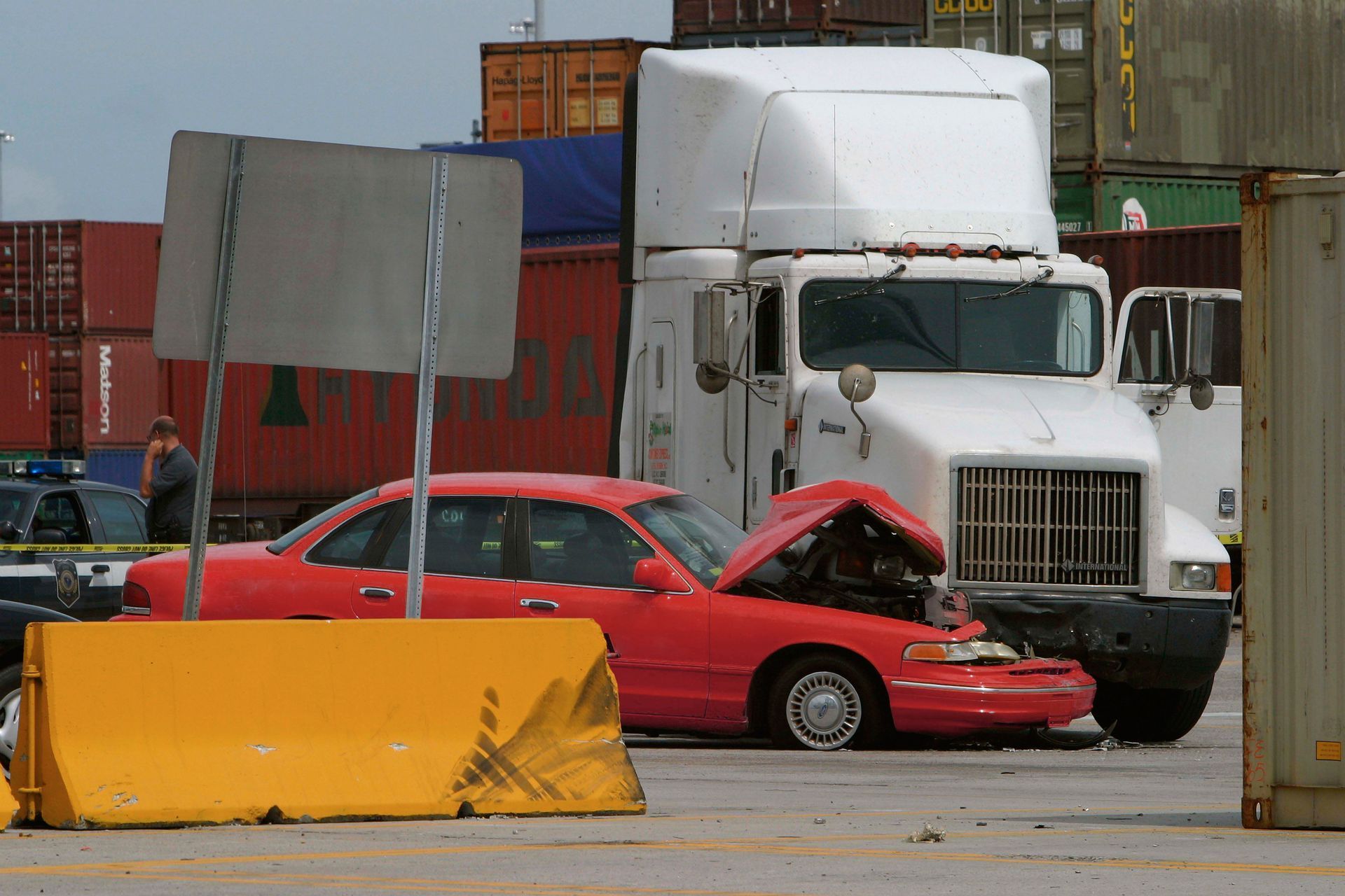 Red car crumpled under a white truck in a shipping yard with containers and barriers