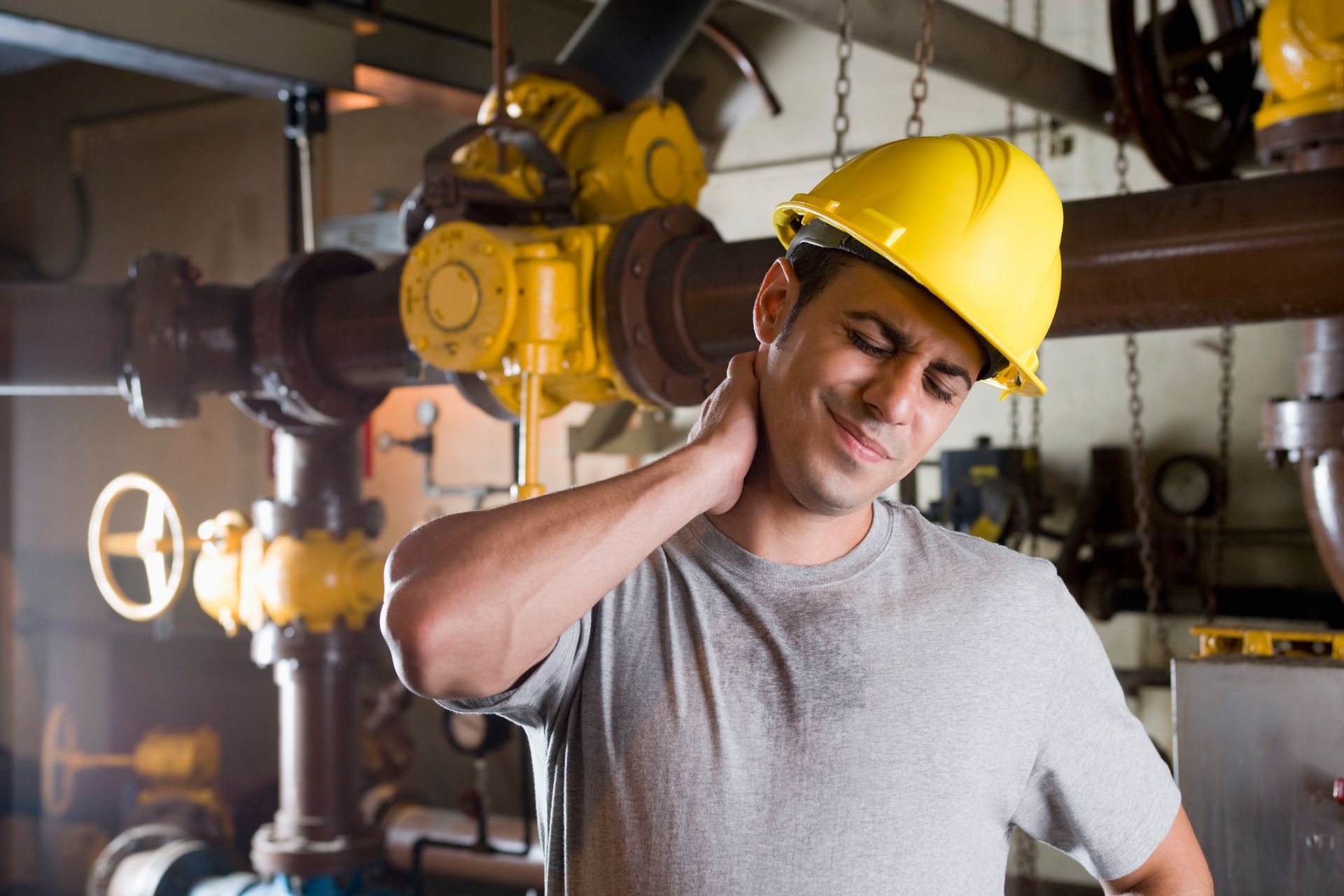 Worker in yellow hard hat rubbing his neck beside industrial pipes and valves.
