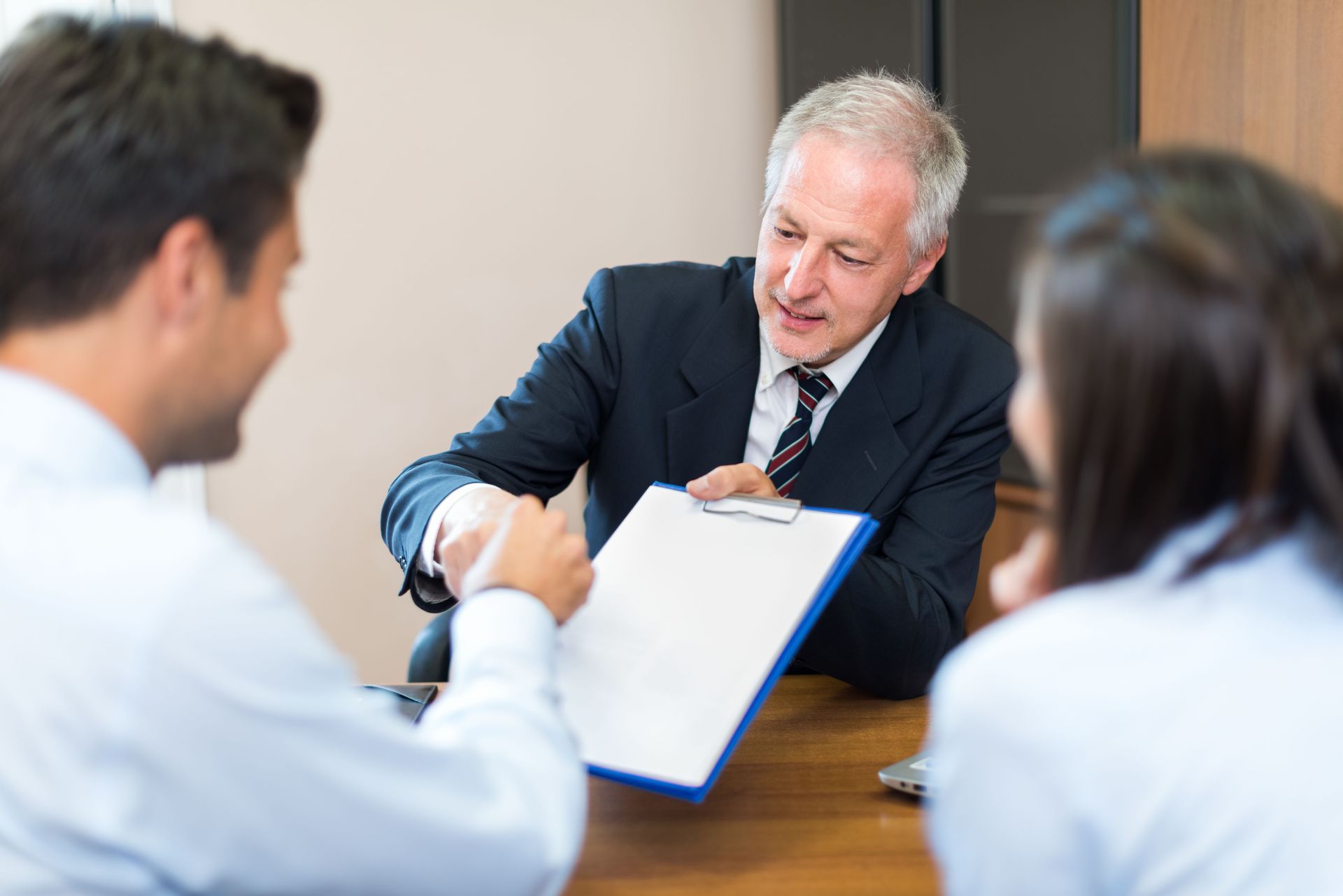 Three people in a business meeting, one taking notes on a clipboard across a table.