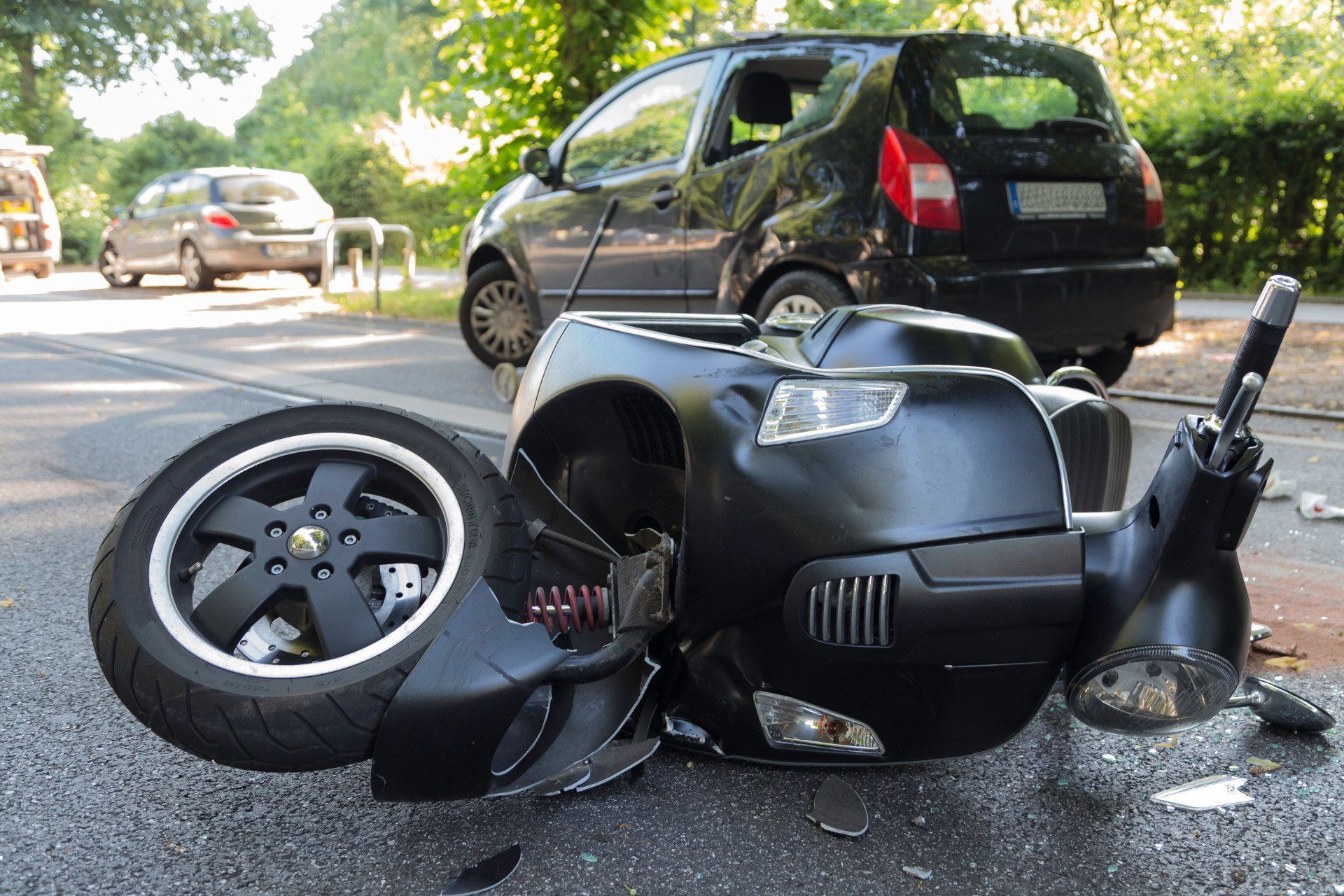 Overturned black scooter after a crash, with a detached wheel on a road behind a parked car.