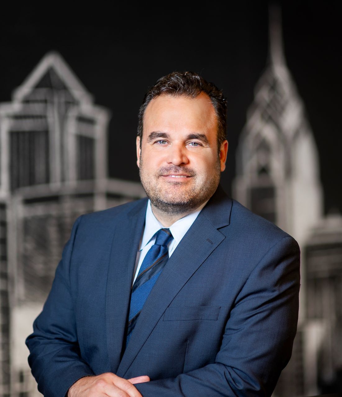 Man in a blue suit standing with arms crossed against a blurred architectural background