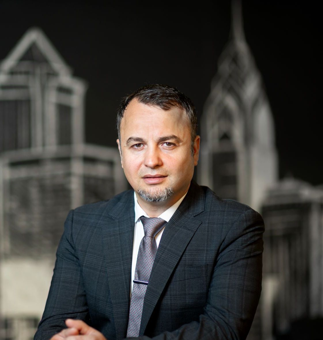 Portrait of a man in a dark suit and tie against a blurred black-and-white architectural backdrop