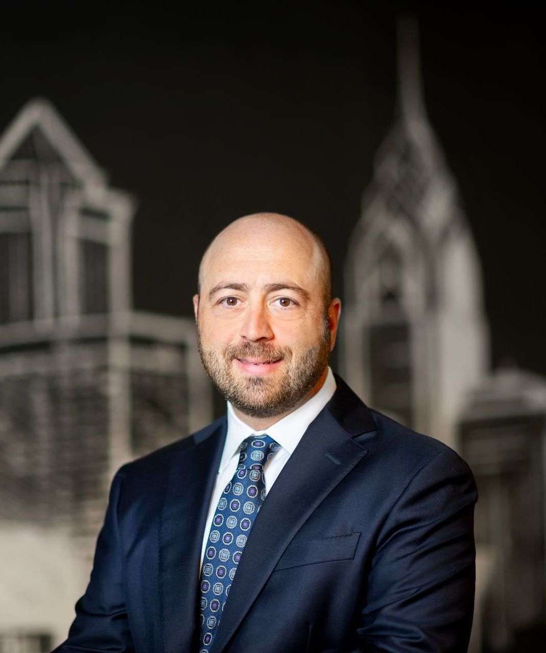 Man in a navy suit and blue tie posing in front of a blurred city skyline backdrop