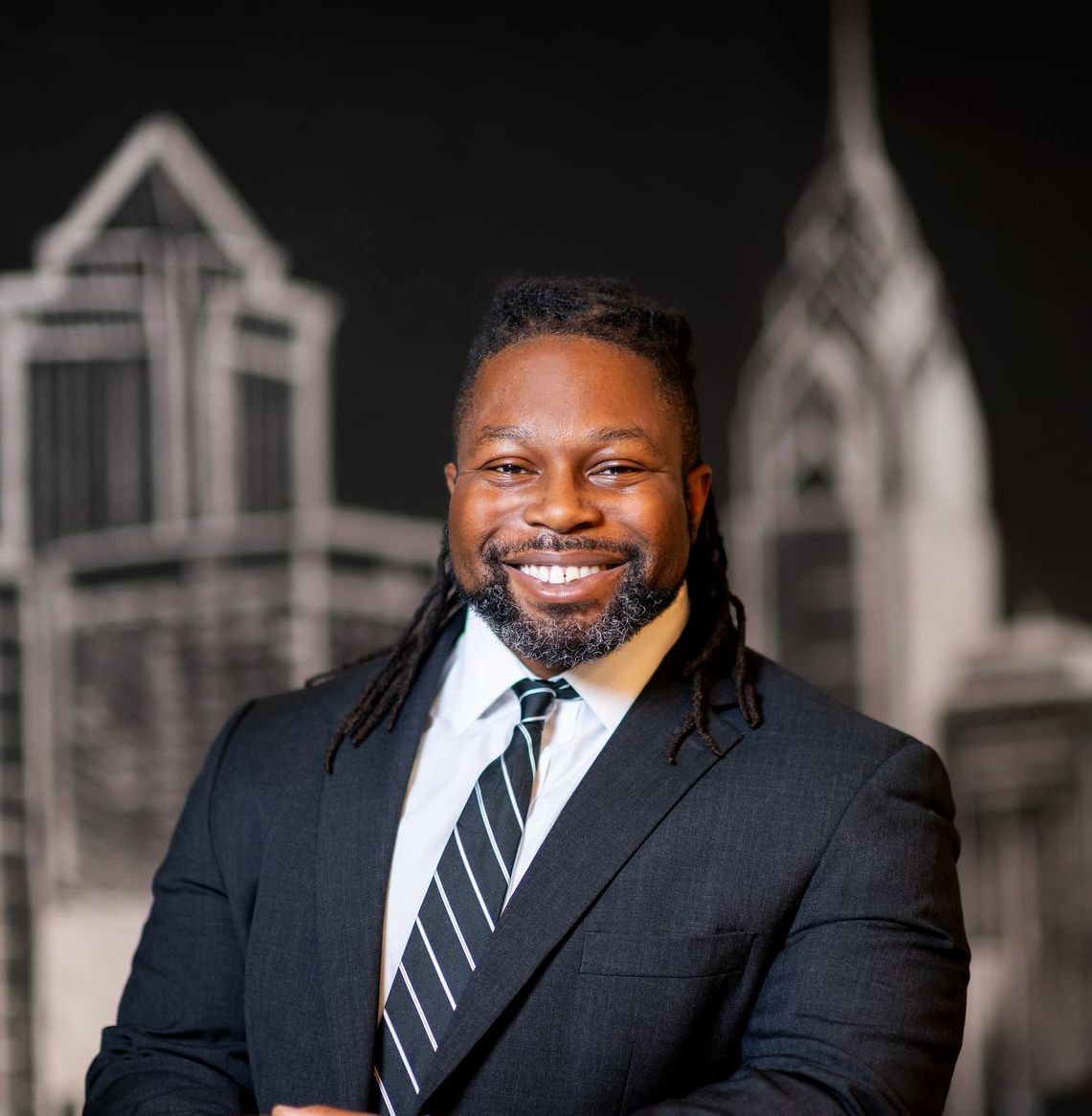 Smiling man in a dark suit and striped tie, standing in front of a blurred architectural backdrop