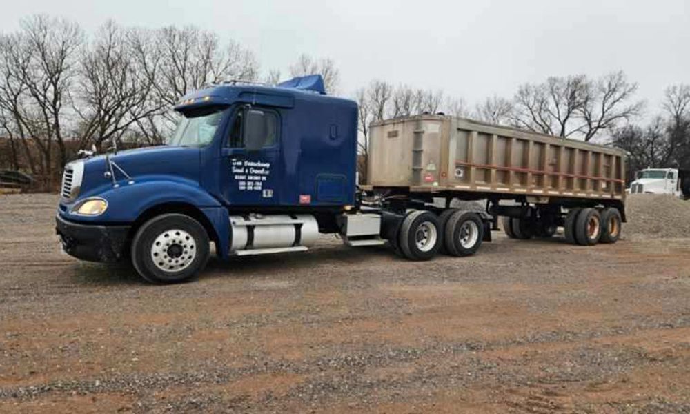 A blue semi truck with a dump trailer attached to it is parked in a dirt lot.
