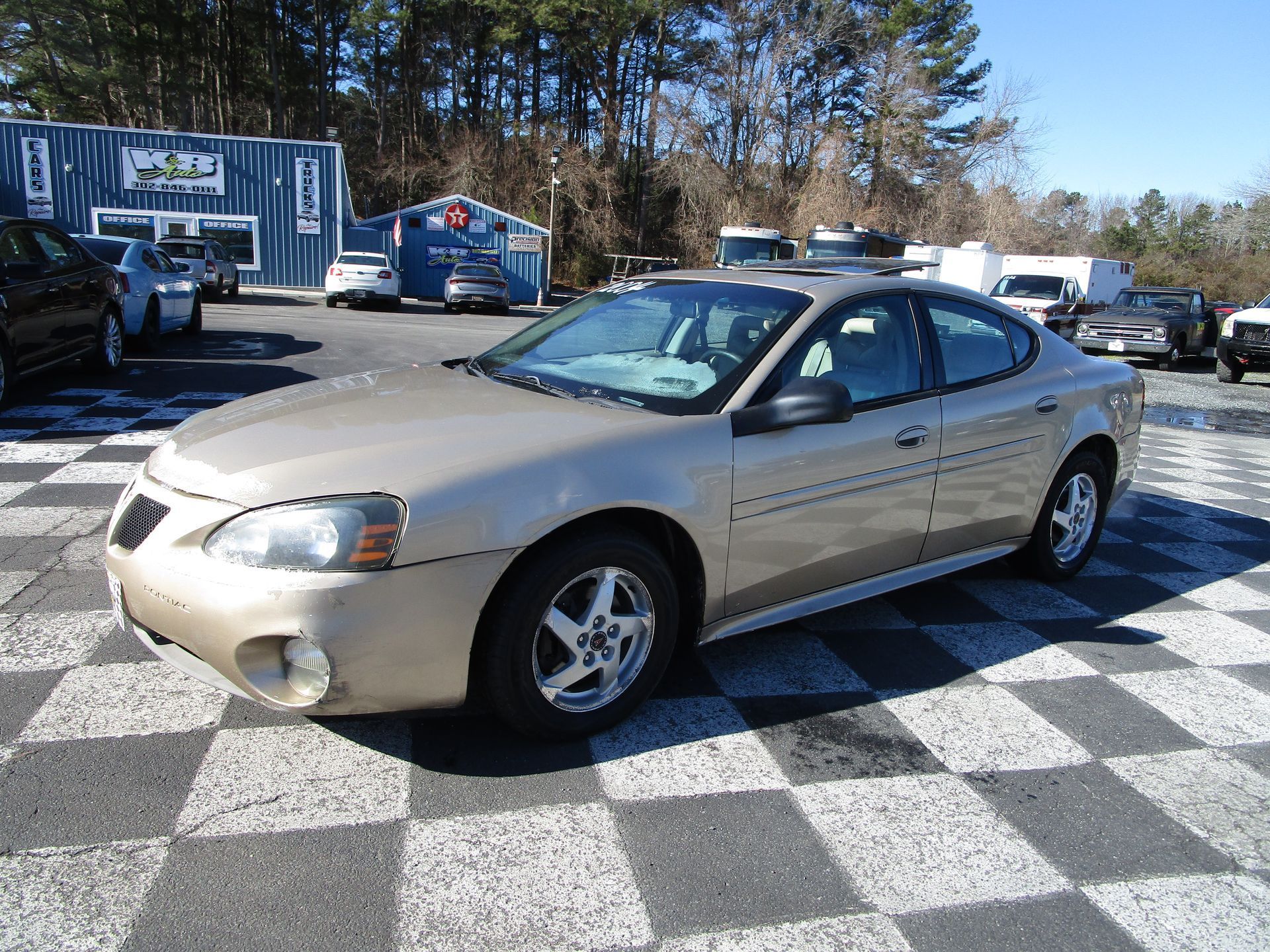 Gold Pontiac Grand Prix sedan parked on a checkered lot at a car dealership on a sunny day.