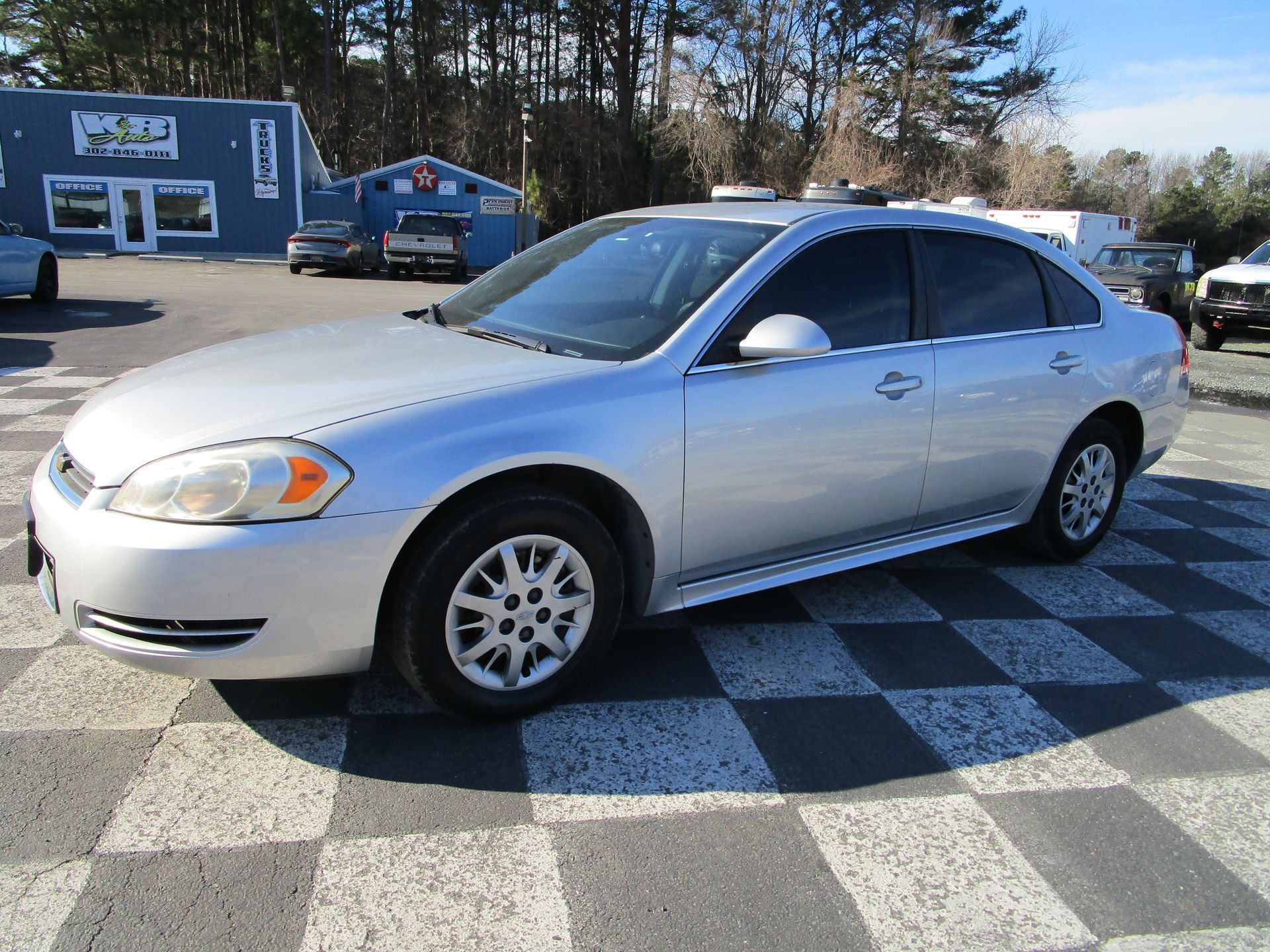 Silver Chevrolet Impala sedan parked on checkered pavement in front of a blue building on a sunny day.