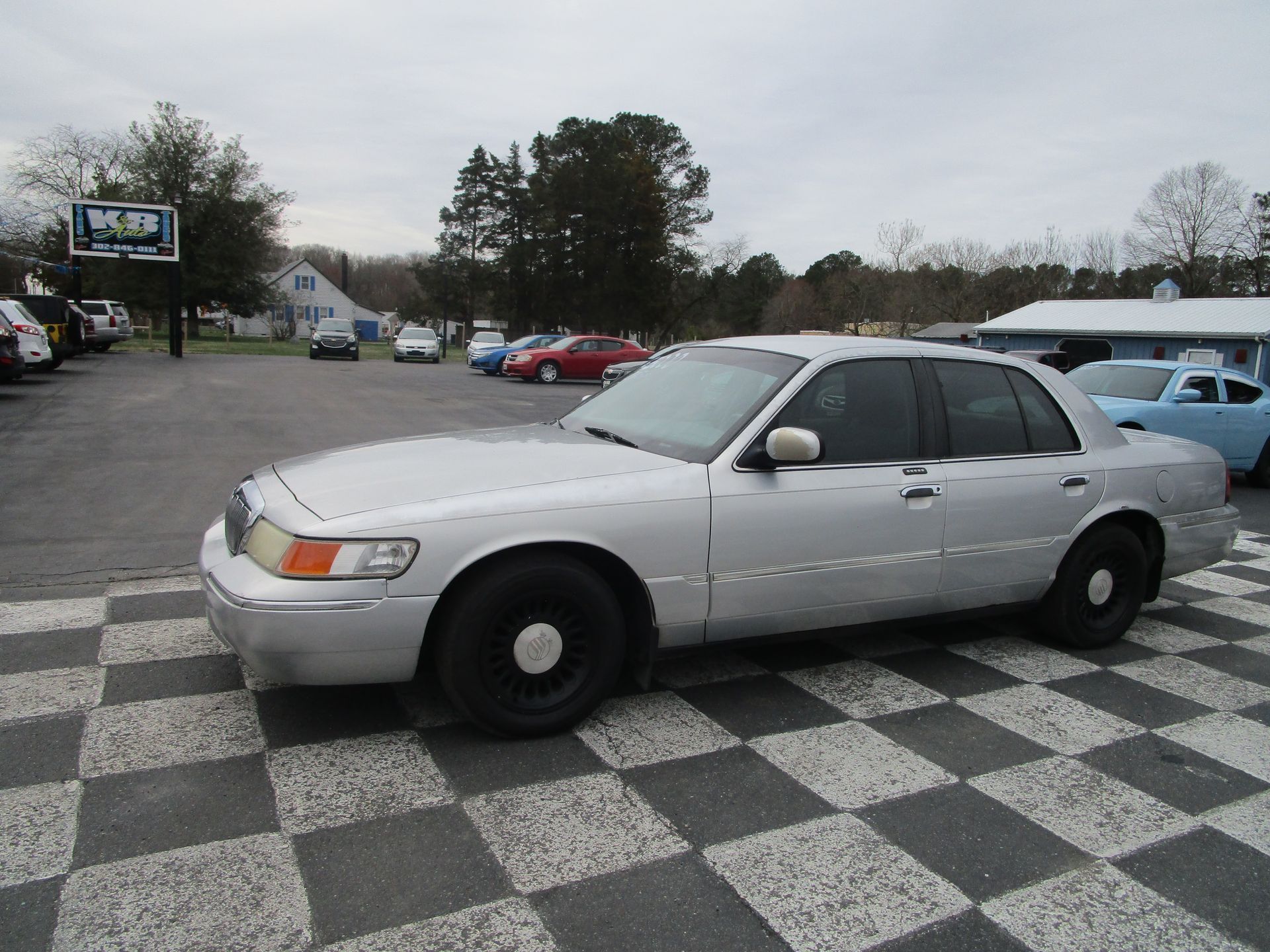 A silver Ford Crown Victoria parked on a checkered lot in front of a car dealership.