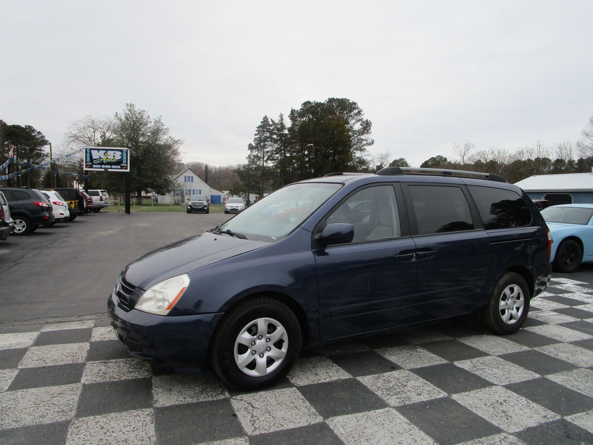 A dark blue minivan parked on a checkered lot at a car dealership on an overcast day.