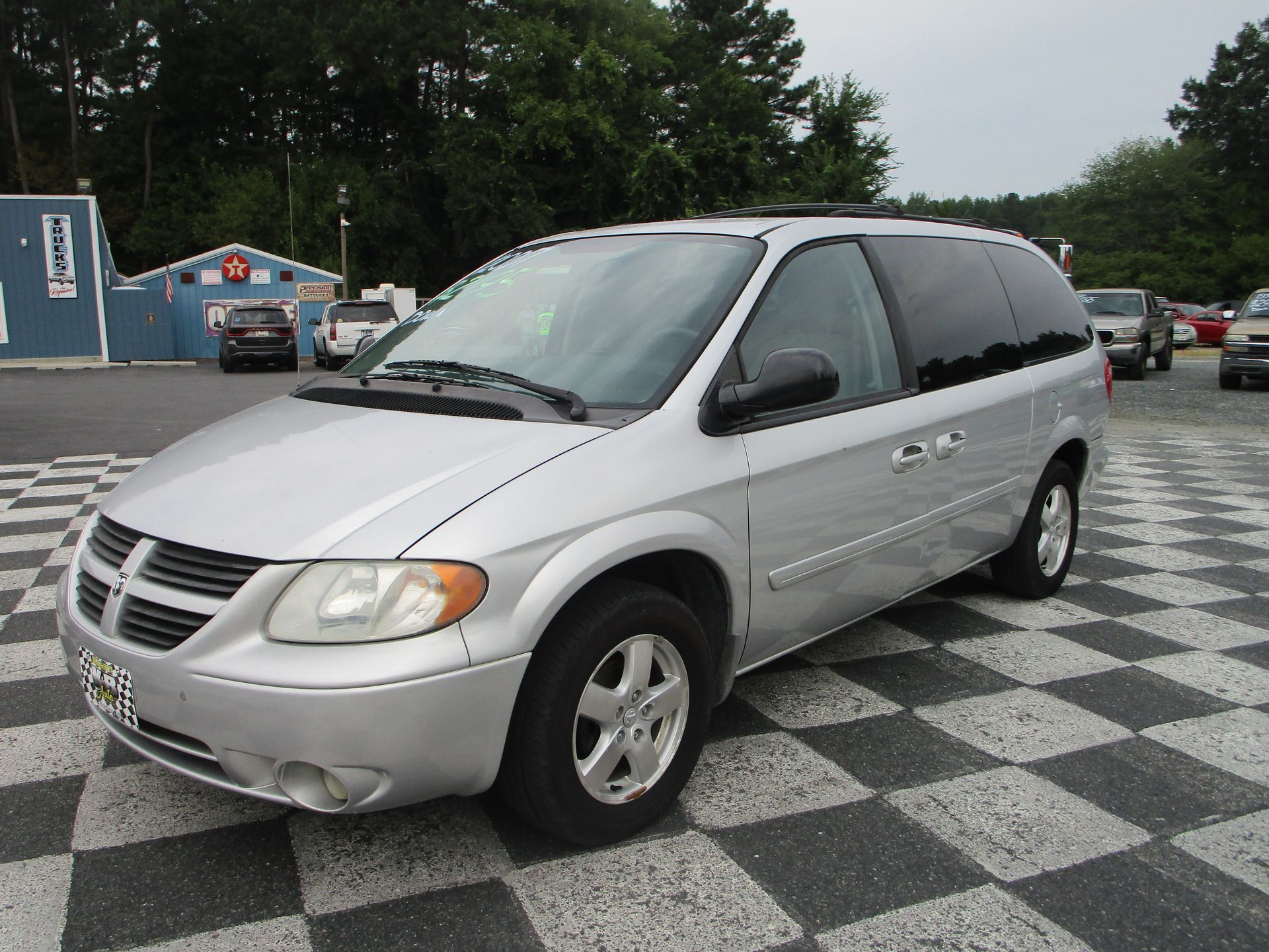 Silver Dodge Caravan minivan parked on a checkered surface, in front of a building with other vehicles visible.