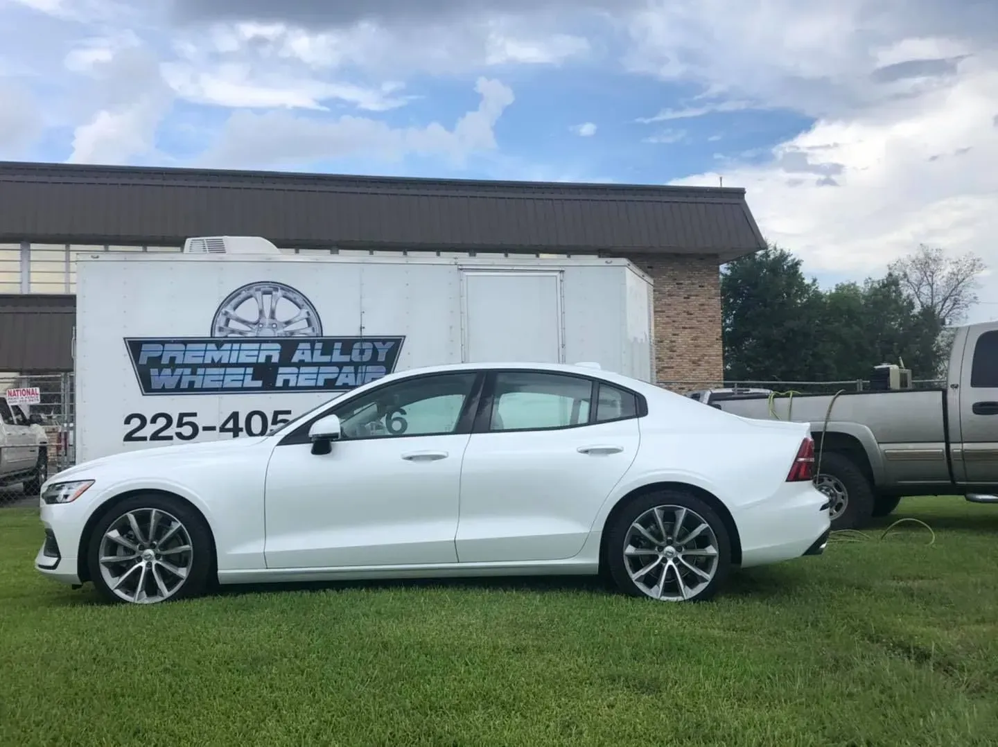 White sedan parked on grass in front of a building and truck trailer under a cloudy sky