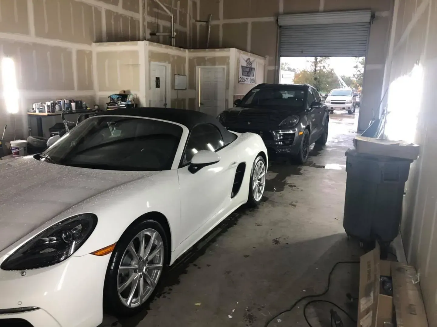White sports car and black SUV parked inside a garage with unfinished walls and a trash bin in the foreground