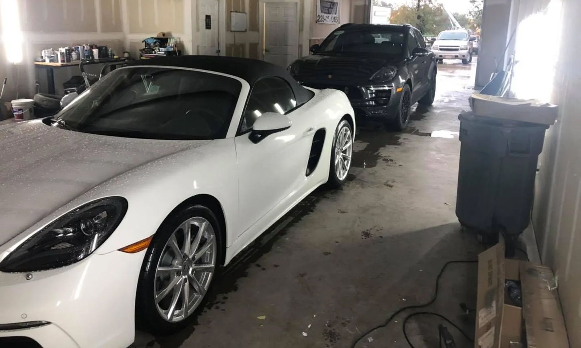 White convertible and black SUV parked in a garage-like carport with a trash bin nearby