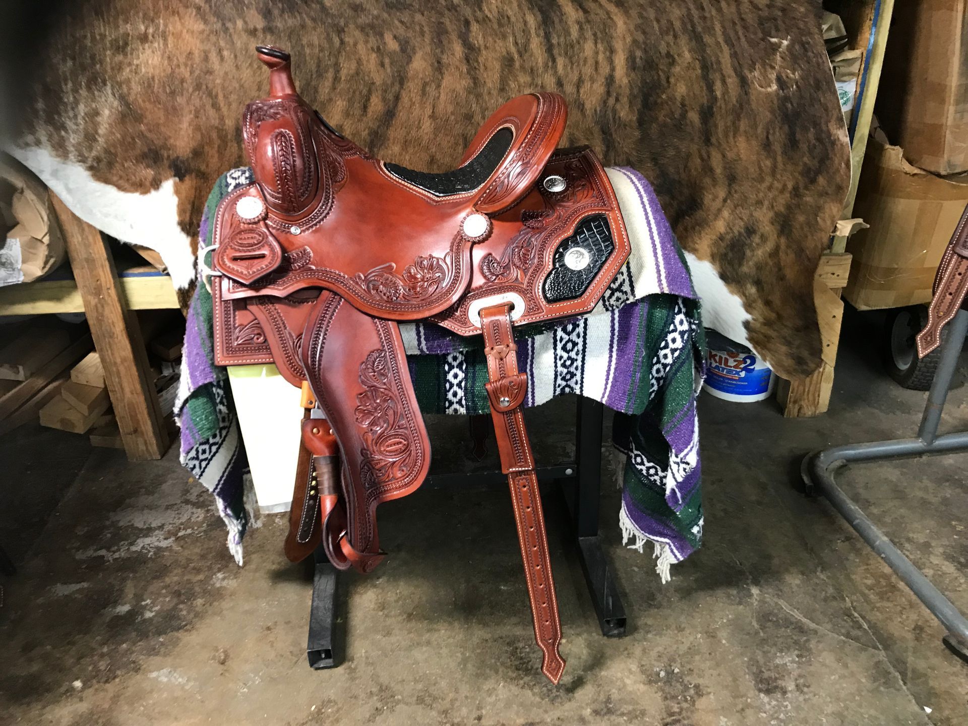 A brown, tooled leather Western saddle rests on a stand covered by a purple, green, and white striped saddle blanket.
