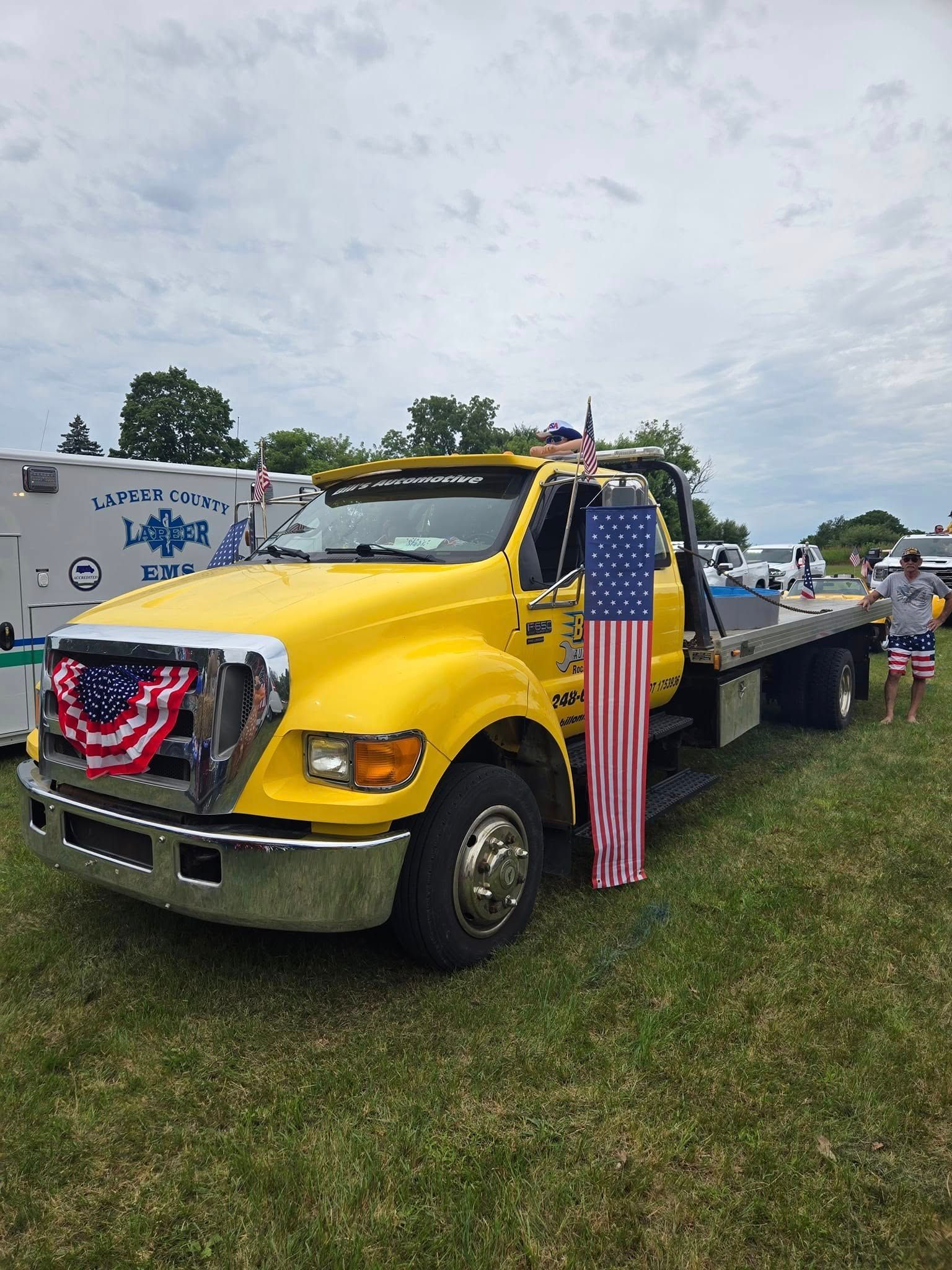 A yellow tow truck with an american flag on the back is parked in a grassy field.