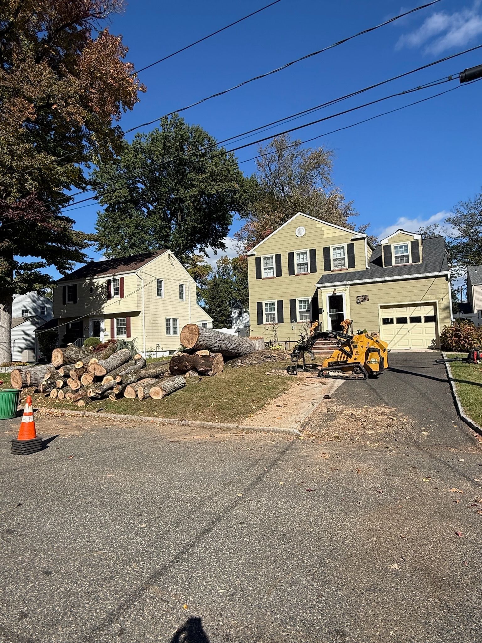 A residential street with tree removal in progress. Logs and equipment are in the yard of a two-story yellow house.