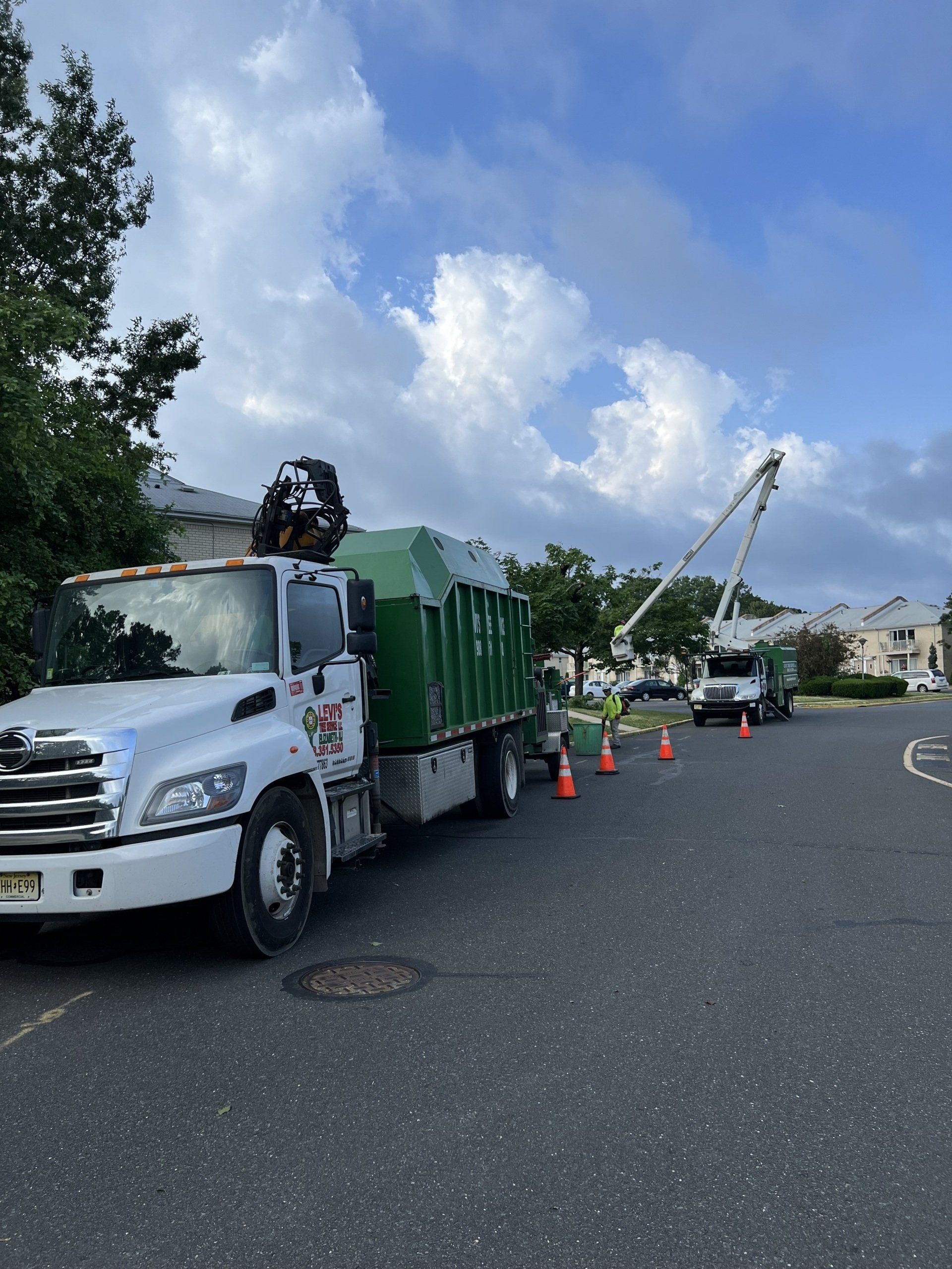 Garbage truck loading a green bin on a street, another truck with a lifted arm in the background, under a cloudy sky.