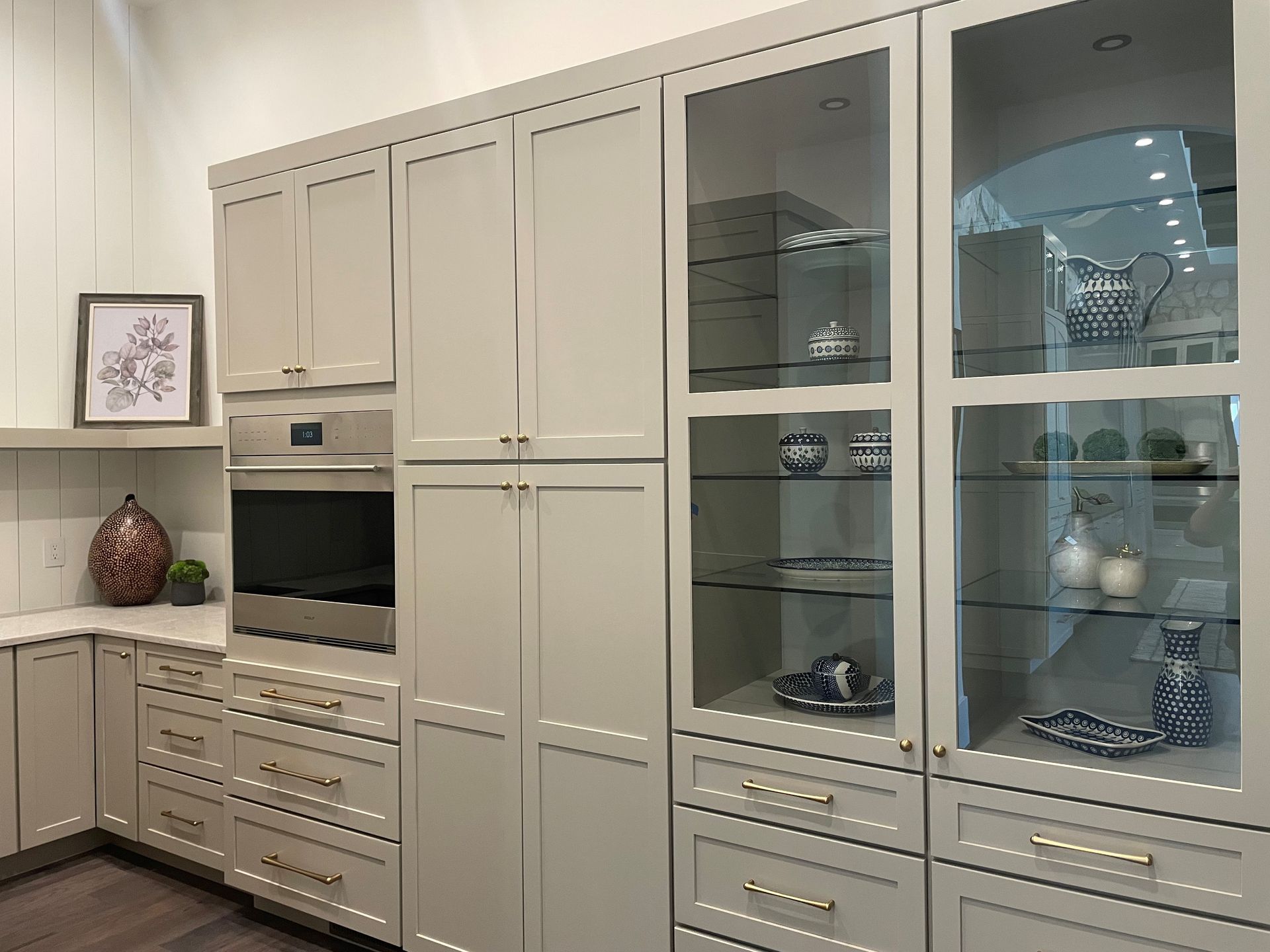 Kitchen cabinets in light gray with glass-front display and built-in oven, featuring gold hardware.