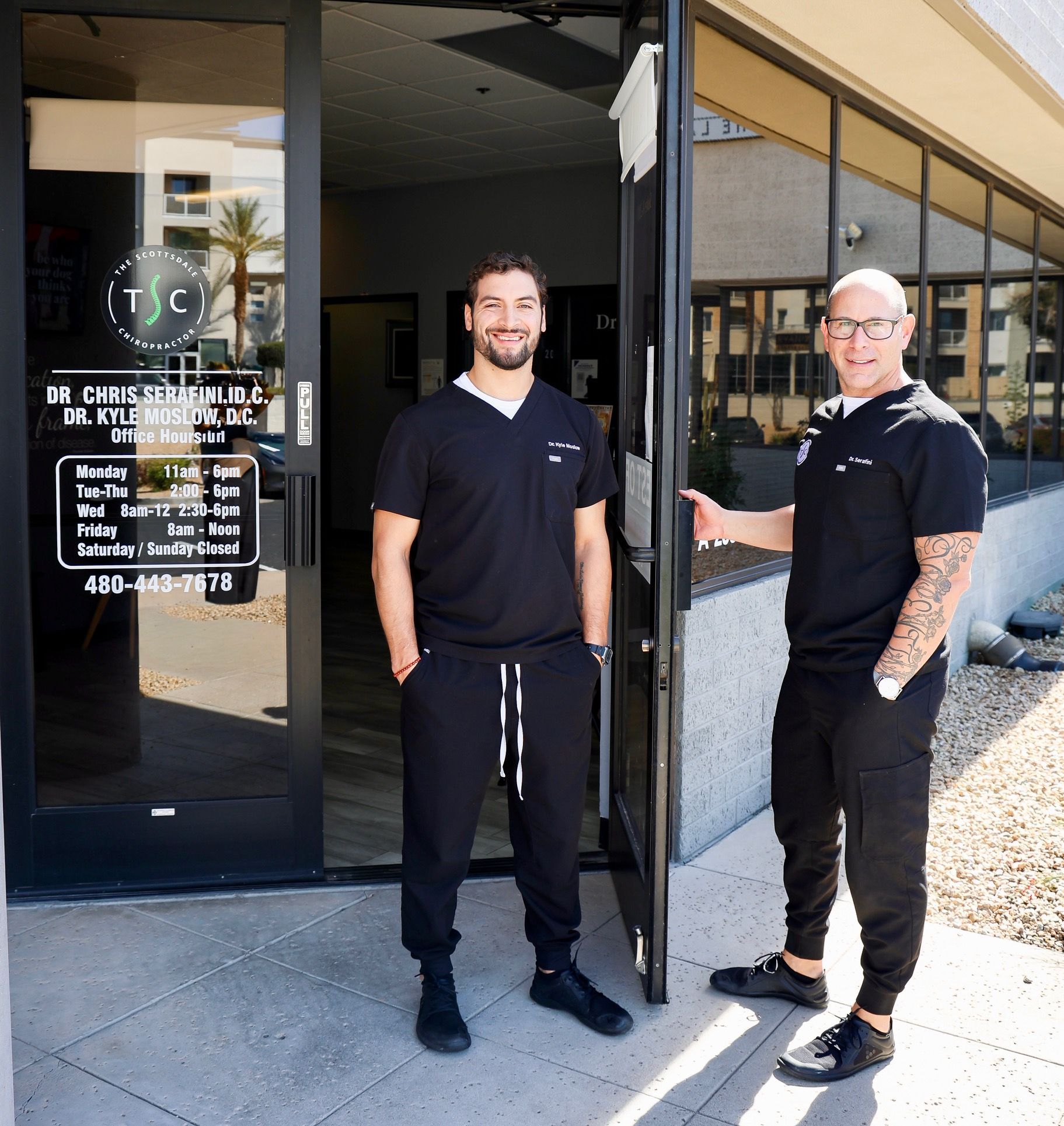 Two professionals in black scrubs stand at the entrance of a medical office, with one man holding the door open.