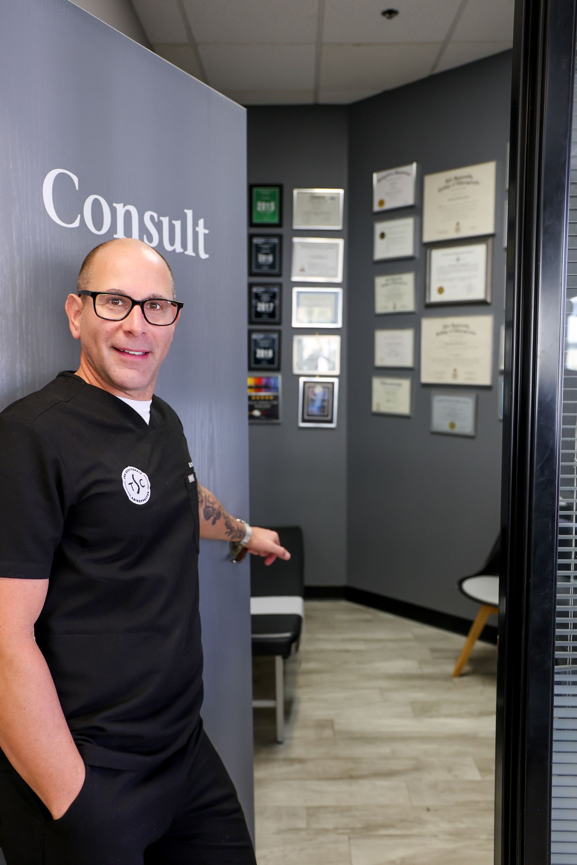 A person in a black scrub top gestures toward a consulting room with walls covered in framed certificates.