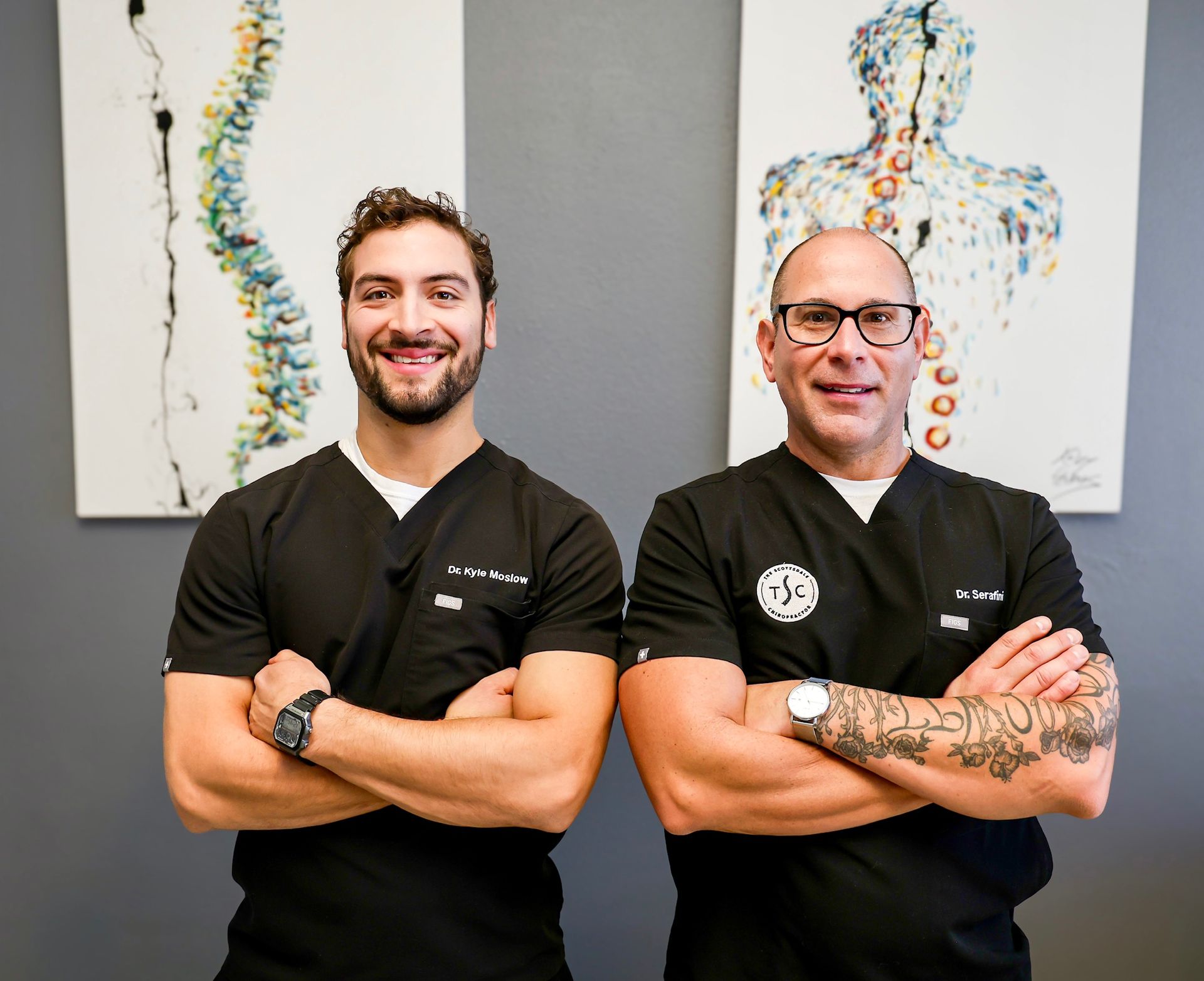 Two people in black medical scrubs stand with arms crossed in front of medical-themed artwork on a gray wall.