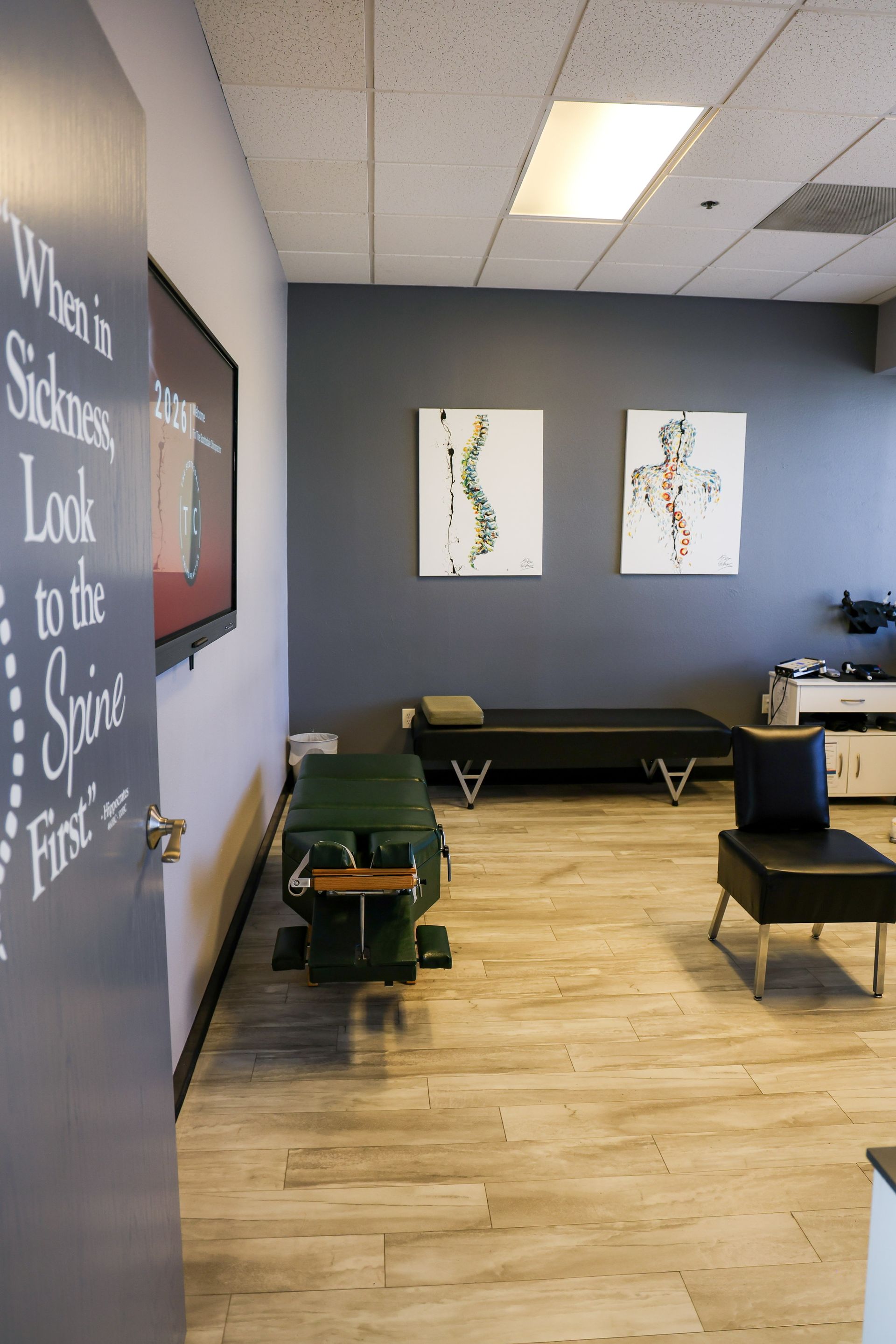 A chiropractic office with a treatment table, chairs, spinal art on a gray wall, and a quote on the door.